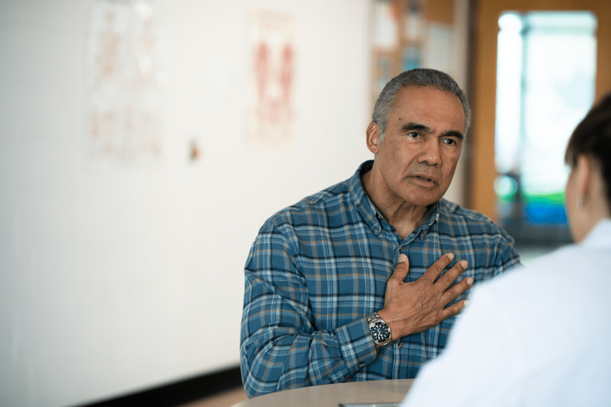 A photograph of two people sitting at a table. Facing us is a man in a long-sleeved blue plaid shirt with his right arm on his chest. He is looking across the table at someone in a lab coat, who has their back to us.